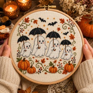 May include: A circular wooden embroidery hoop containing a cross-stitch pattern on cream fabric. The design features four white ghosts holding black umbrellas, surrounded by orange pumpkins, autumn leaves, green vines, and small black bats.
