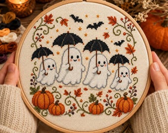 May include: A circular wooden embroidery hoop containing a cross-stitch pattern on cream fabric. The design features four white ghosts holding black umbrellas, surrounded by orange pumpkins, autumn leaves, green vines, and small black bats.