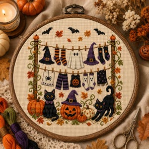 May include: A circular wooden embroidery hoop containing a cross-stitch design of Halloween items hanging on a clothesline, including witch hats, socks, a ghost, and a towel. Two black cats and pumpkins are at the bottom with autumn leaves.