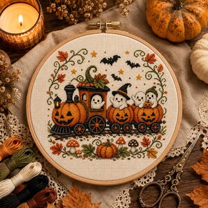 May include: A round wooden embroidery hoop containing a cross-stitch pattern of a pumpkin train. The train features three orange jack-o'-lantern cars carrying white ghosts, with one ghost wearing a witch hat. The design includes bats, stars, autumn leaves, and mushrooms.