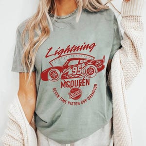 May include: A light green t-shirt featuring a red graphic of a race car with the text "Lightning McQueen 95" and "Seven Time Piston Cup Champion". The shirt has a vintage, worn look.