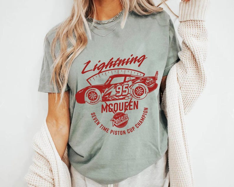 May include: A light green t-shirt featuring a red graphic of a race car with the text "Lightning McQueen 95" and "Seven Time Piston Cup Champion". The shirt has a vintage, worn look.