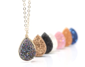 May include: A gold chain necklace with a teardrop-shaped pendant made of iridescent purple, blue, and gold druzy. The pendant is surrounded by a gold wire. The necklace is displayed with other teardrop-shaped pendants in various colors.