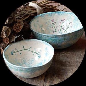 Könnte beinhalten: Zwei Keramikschalen mit hellblauem und weißem Sprenkelmuster. Die Schalen weisen handgemalte Blumenzweige in Blau- und Lilatönen auf. Die Schalen stehen auf einer Holzoberfläche.