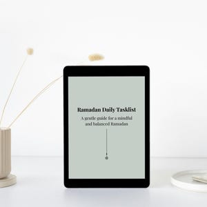 May include: A black tablet displays the text "Ramadan Daily Tasklist" on a light green background. The text is in black, with a vertical line and a small circle below. A beige vase with dried flowers and a white tray with a silver clip are also in the image.