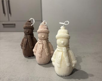 May include: Three snowman-shaped candles in varying shades of brown, pink, and white. Each candle features a knitted hat and scarf, with a looped wick at the top. The candles are arranged in a row, showcasing their winter theme.