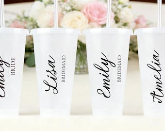 May include: Four white plastic tumblers with lids and straws, each with a name in elegant black script. One says "Emily Bride", the others say "Lisa Bridesmaid", "Emily Bridesmaid", and "Amelia Bridesmaid". A floral arrangement is in the background.