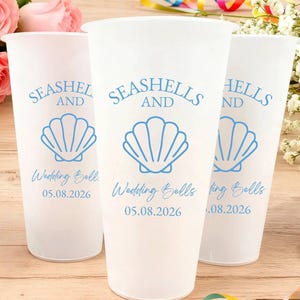 May include: Three white plastic cups with blue text and seashell graphics. The text reads "Seashells and Wedding Bells 05.08.2026". The cups are on a wooden surface with flowers and ribbons.