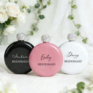 May include: Three circular flasks with crystal-embellished stoppers. One is black with "Amber BRIDESMAID" in white script, another is pink with "Emily BRIDESMAID", and the third is white with "Daisy BRIDESMAID". The flasks are displayed in front of white flowers.