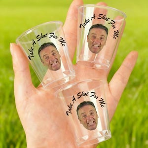 May include: Four clear plastic shot glasses, each with a smiling man's face and the text "Take A Shot For Me" in black script. The glasses are held in a hand against a blurred green background.