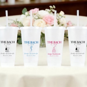 Puede incluir: Cuatro vasos de plástico transparente con tapas y pajitas, cada uno impreso con "THE BACH Club" y "Emily's Bachelorette March 9, 2026" en varios colores. Los vasos están sobre un mantel blanco, con flores en el fondo.