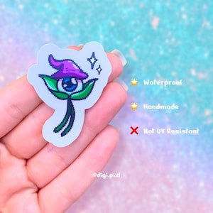 Peut inclure: Un autocollant pixelisé avec un motif d'œil fantaisiste, orné d'un chapeau violet et de feuilles vertes. L'autocollant est étiqueté "Waterproof" et "Handmade", avec une note "Not UV Resistant". Le sticker a une bordure blanche.