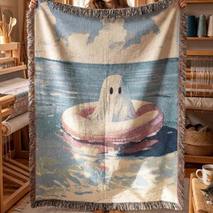Può includere: Una coperta tessuta raffigurante un fantasma sotto un lenzuolo che galleggia in un salvagente rosa su acqua blu sotto un cielo nuvoloso. La coperta rettangolare ha bordi sfrangiati e mostra una scena dettagliata di un fantasma nell'oceano.