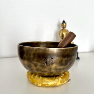 Puede incluir: Un cuenco tibetano de bronce con un diseño tallado, sobre un cojín amarillo. Una maza de madera descansa dentro del cuenco. Una pequeña estatua dorada de Buda está en el fondo.