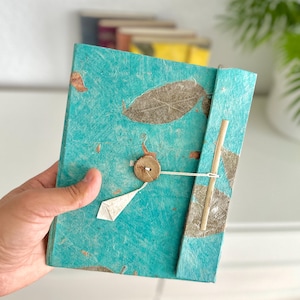 Op de afbeelding: Een handgemaakt notitieboek met een turquoise omslag met ingebedde bladeren en een houten knoopsluiting. Het notitieboek wordt opengehouden en onthult het getextureerde papier en de natuurlijke elementen die in het ontwerp zijn gebruikt. Andere notitieboeken zijn zichtbaar op de achtergrond.