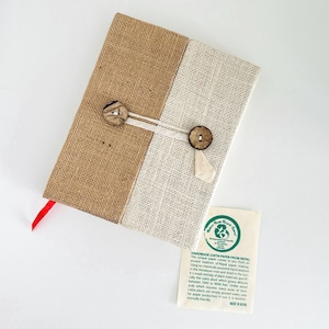 Könnte beinhalten: Ein handgefertigtes Lokta-Papier-Journal aus Nepal. Das Journal hat einen Bezug aus Jute und cremefarbenem Stoff mit Holzknopfverschluss. Ein rotes Band-Lesezeichen ist sichtbar. Eine kleine Karte mit Text über das Papier ist enthalten.