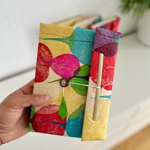 Op de afbeelding: Een handgemaakt notitieboek met een kleurrijk bladerenontwerp. De omslag heeft een patroon van rode, roze, blauwe en groene bladeren op een crèmekleurige achtergrond. Het is vastgemaakt met een knoop- en touwsluiting.