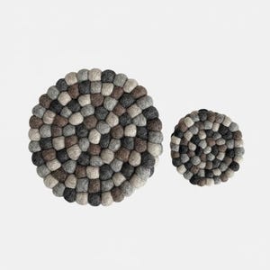 May include: Two circular felt trivets in various shades of grey, brown, and beige. The trivets are constructed from small, densely packed felt balls, providing a textured surface. One trivet is larger than the other, both set against a white backdrop.