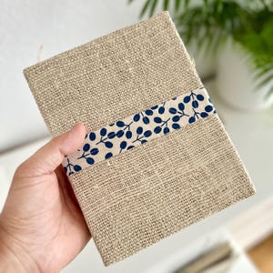 Può includere: Un diario rettangolare con una copertina in juta naturale. Una fascia a motivi blu e bianchi con disegni di foglie avvolge il centro. Il diario è tenuto in mano, suggerendo un prodotto artigianale.
