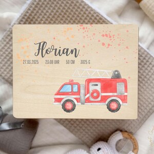 以下が含まれることがあります： 赤い消防車のイラストが描かれた木製プレート。名前の「Florian」は筆記体で書かれており、日付、時間、長さ（50cm）、重さ（3025g）も記載されています。背景にはオレンジとピンクのペンキの飛沫があります。