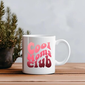 May include: White ceramic mug with the text "Cool Moms Club" in a gradient of pink and red. The mug is on a wooden surface with a small potted plant in the background.