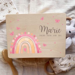 以下が含まれることがあります： 虹と蝶のデザインが施された明るい木製の箱。箱には「Marie」という名前が書かれており、日付、時間、長さ（50cm）、重さ（3150g）も記載されています。デザインにはピンクとオレンジ色が使用されています。
