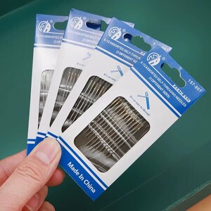 Könnte beinhalten: Vier Packungen selbstfädelnder Nadeln werden in einer Hand gehalten. Jede Packung ist weiß und blau, mit dem Text "6/12 ASSORTED SELF-THREADING NEEDLES" und "Made In China". Die Nadeln sind durch ein klares Fenster sichtbar.