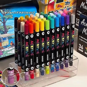 May include: A set of acrylic markers in various colors, displayed in a clear plastic organizer. The markers have black barrels with the word "ACRYLIC MARKER" printed on them. The caps and tips are color-coded. The image also shows a box with the text "12 Acrylic Markers".