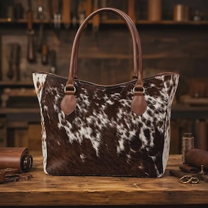 May include: A tote bag featuring a brown and white cowhide pattern with smooth brown leather handles and trim. The bag has a wide opening, a structured base, and gold-toned metal hardware connecting the handles to the main body of the bag.