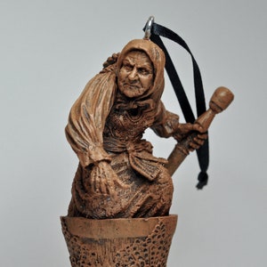 May include: A brown ceramic ornament of a woman holding a staff, standing in a brown ceramic cup with a decorative pattern. The ornament has a black ribbon for hanging.