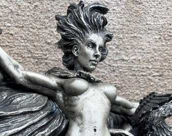 May include: A silver-toned sculpture of a woman with flowing hair and wings, holding a raven in each hand. The woman is standing on a base with a swirling design.