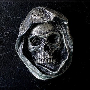 May include: A silver skull with a hooded cloak, the skull has a detailed design and is a decorative piece.