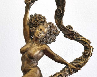 May include: Bronze sculpture of a nude woman holding a floral vine. The sculpture has a dark brown base with floral and rock details. The woman's hair is curly, and the sculpture has a classical aesthetic.