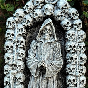 May include: A gray, stone-like sculpture featuring a hooded figure in a long robe standing within an archway constructed of stacked human skulls. The figure has skeletal hands clasped together, and the base is textured like stone masonry blocks.