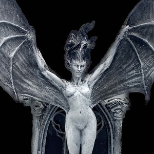 Pode incluir: Uma escultura de uma figura feminina alada branca com um arco g&oacute;tico atr&aacute;s. A escultura &eacute; feita de um material texturizado e tem um aspecto desgastado. A figura est&aacute; em p&eacute; em um conjunto de degraus com dois casti&ccedil;ais de cada lado.