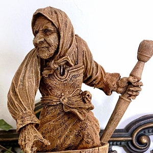 May include: A wooden figurine of a woman holding a wooden spoon, standing in a wooden mortar and pestle. The figurine is painted in shades of brown and has a detailed face.