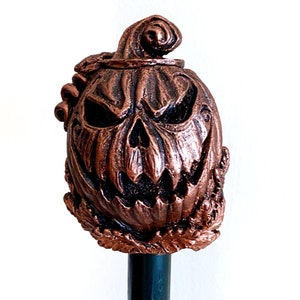 May include: A copper-colored Halloween pumpkin head with a carved scary face, attached to a black wooden stick.