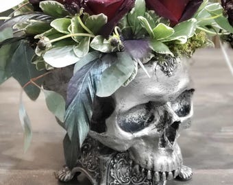 May include: A silver skull-shaped vase filled with deep red roses and various green and purple foliage. The vase has intricate details and sits on a wooden surface, creating a gothic floral arrangement.