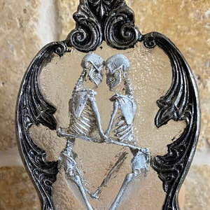 May include: A silver and black nightlight with a glass insert featuring two skeletons dancing. The skeletons are white and the glass is frosted. The nightlight is mounted on a wooden base.