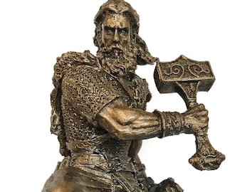 May include: A bronze-colored hanging ornament depicting a bearded warrior in chainmail armor, leaping forward while wielding a large hammer with intricate scrollwork on the head. The figure wears wrapped leg coverings and a belt, suspended by a loop.
