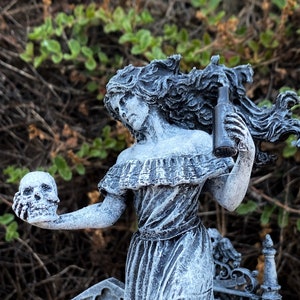May include: A gray statue of a woman holding a skull and a bottle, with a graveyard scene in the background. The woman is wearing a dress and has long, flowing hair. The statue is detailed with a weathered finish, evoking a gothic theme.