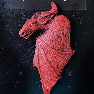 May include: A red dragon sculpture with detailed wings and a textured surface. The dragon is in a curled position with its head turned to the side.
