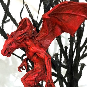 Op de afbeelding: Rode demonen ornament met vleugels en een lange staart, hangend aan een kale tak.