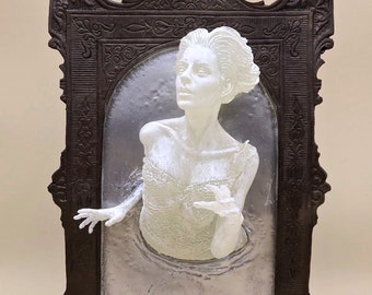 May include: A white figurine of a woman emerging from a silver frame. The frame is ornate and dark brown with intricate details. The woman is wearing a white dress and has a distressed look on her face.