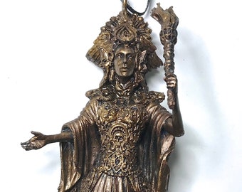 May include: A bronze-colored figurine of a woman wearing a crown and a long flowing robe. She is holding a torch in her right hand and her left hand is outstretched. The figurine is detailed with intricate designs and patterns.