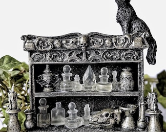 Puede incluir: Una estantería de estilo gótico con una figura de gato negro en la parte superior. La estantería está llena de pequeñas botellas de vidrio y una calavera. La estantería es gris y tiene una base de tronco de árbol. Hojas verdes y velas a los lados.