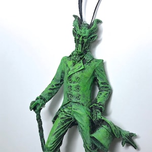 May include: Green Christmas ornament depicting a devilish gentleman in a top hat and coat, holding a cane. The ornament has a black string for hanging.