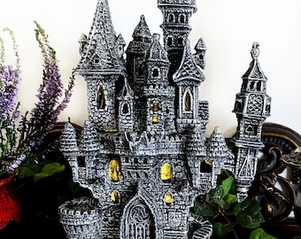 Puede incluir: Una figurita de castillo de piedra gris con un diseño detallado, que incluye torres, ventanas y una puerta. El castillo está sentado sobre una superficie de madera oscura con follaje verde.