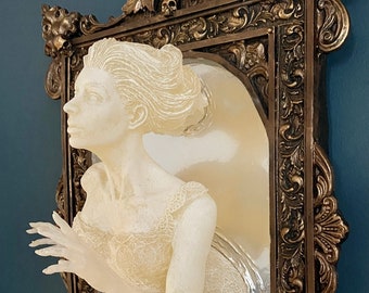 May include: A white, translucent sculpture of a woman's bust emerging from a gold ornate mirror frame. The sculpture is detailed with a lace bodice and a flowing hairstyle. The mirror is set against a dark blue wall.