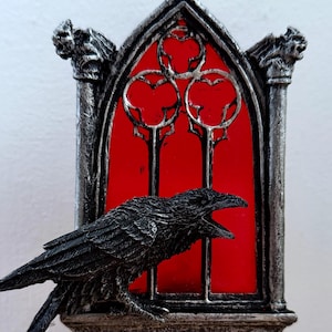Puede incluir: Una luz de noche decorativa con un cuervo negro frente a un diseño de ventana de vidrieras rojas. La ventana está enmarcada en un arco gótico plateado. La luz de noche se enchufa a una toma de corriente estándar.